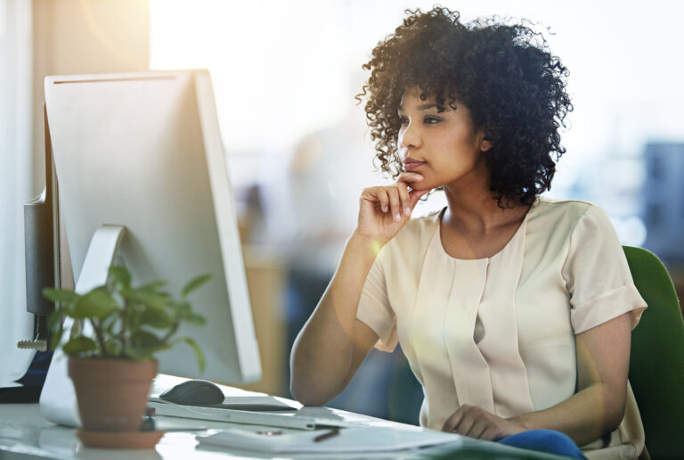 Femme portant un chemisier beige assise à réfléchir devant un écran. Une petite plante repose sur le bureau