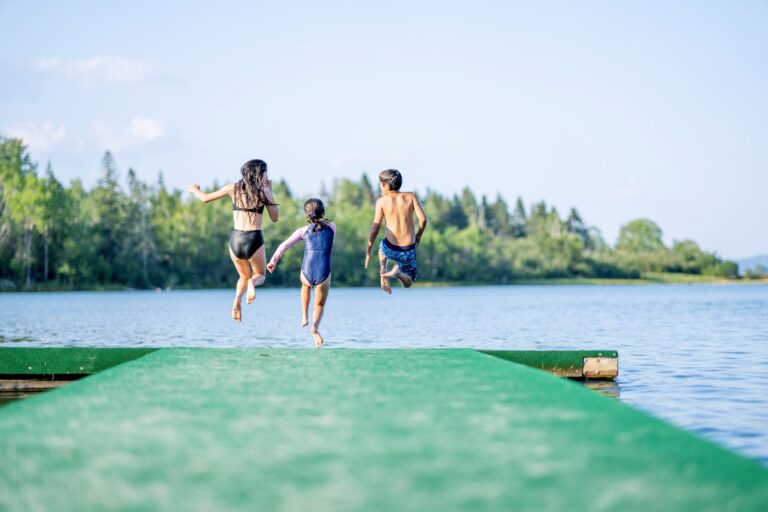 A small group of three children are seen in mid air jumping off the end of a dock on a sunny summer day.
