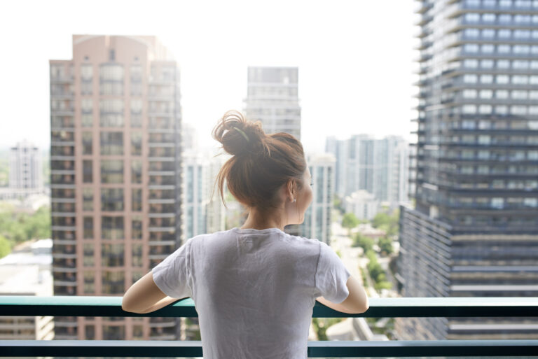 Jeune femme aux cheveux en chignon, debout sur le balcon de sa copropriété, regardant la ville