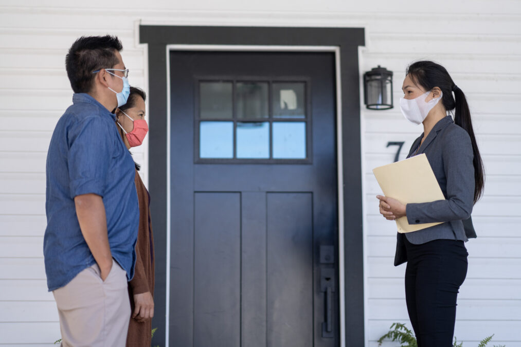 Courtière immobilière se tenant à distance d’un couple de clients d’âge mûr à l’extérieur d’une maison; tous portent un masque