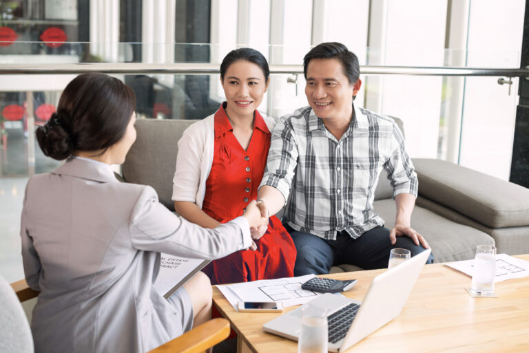 Couple asiatique, femme en robe rouge, assise sur un canapé, souriant et serrant la main d’un courtier immobilier