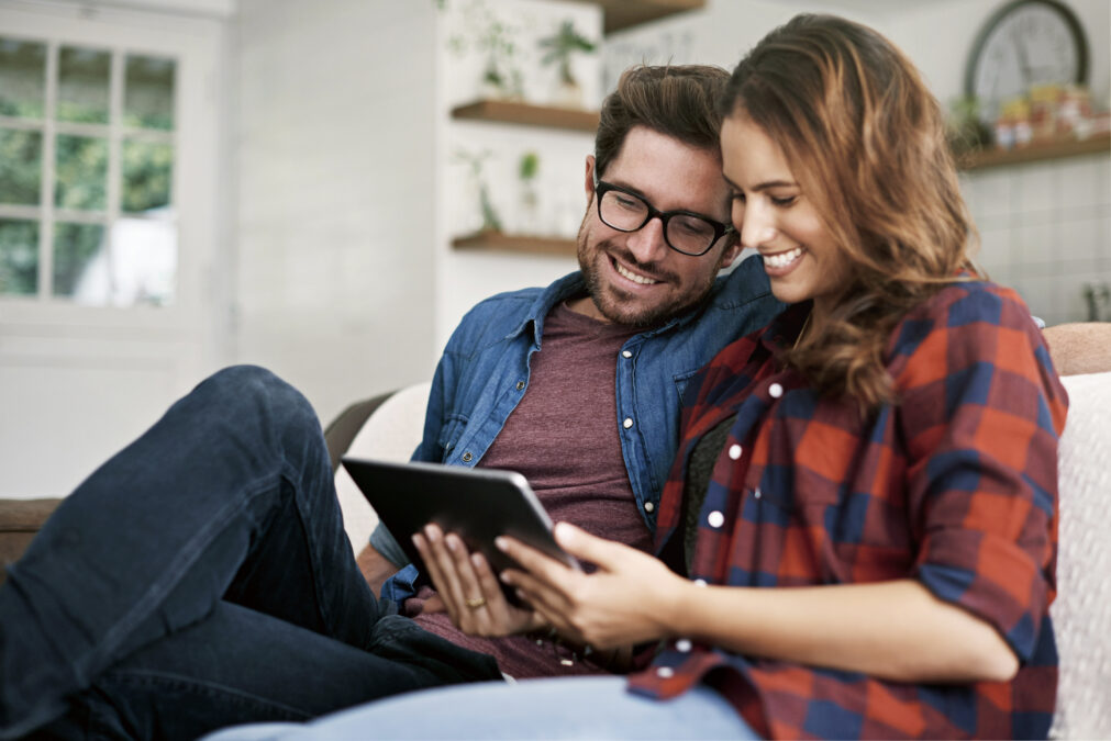Jeune couple souriant, assis sur un canapé, lisant un article sur une tablette, homme en chemise bleue, femme en chemise à carreaux