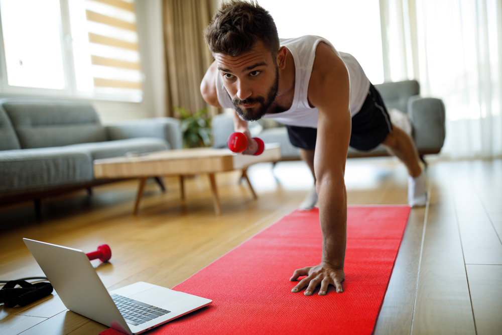 Un homme athlétique en débardeur blanc et en shorts noirs tenant une planche à une main sur un tapis de yoga dans son salon