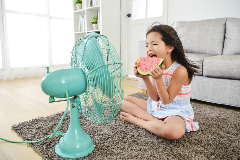 Jeune fille mangeant gaiement du melon d’eau assise devant un ventilateur bleu, se rafraîchissant par une chaude journée d’été