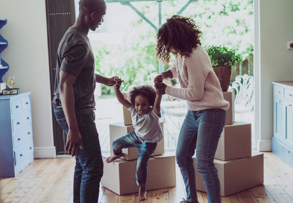 Des parents jouent avec un jeune enfant dans leur salon, avec des boîtes de déménagement et une grande fenêtre donnant sur la cour en arrière-plan