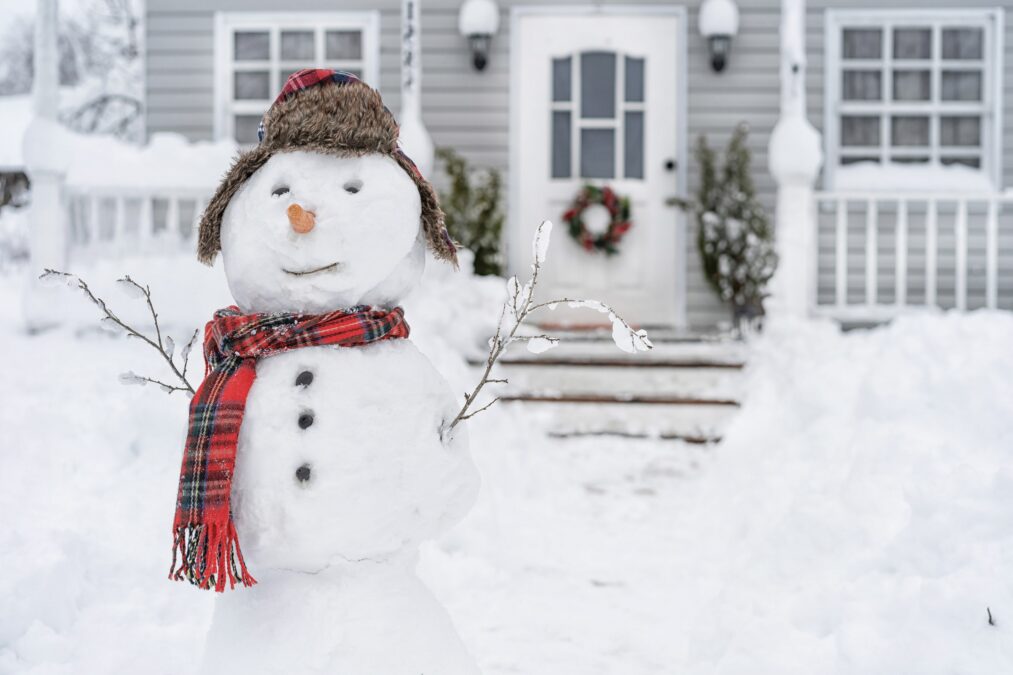 Bonhomme de neige vêtu d’un chapeau et d’un foulard à carreaux devant une maison à l’hiver