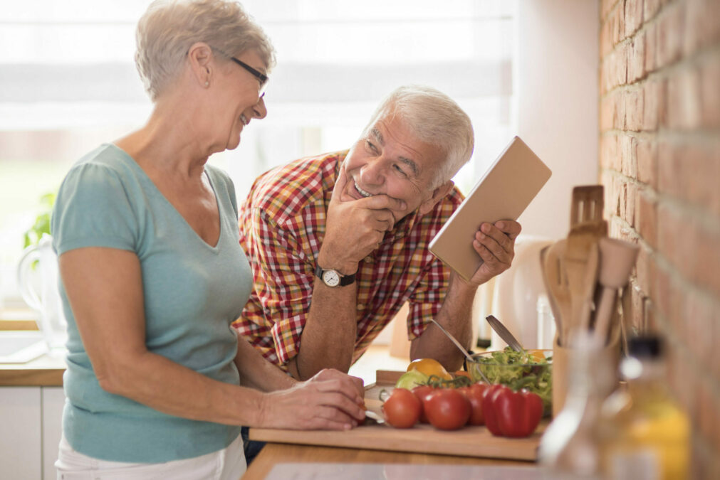 Couple âgé riant et souriant dans sa cuisine tout en préparant un repas