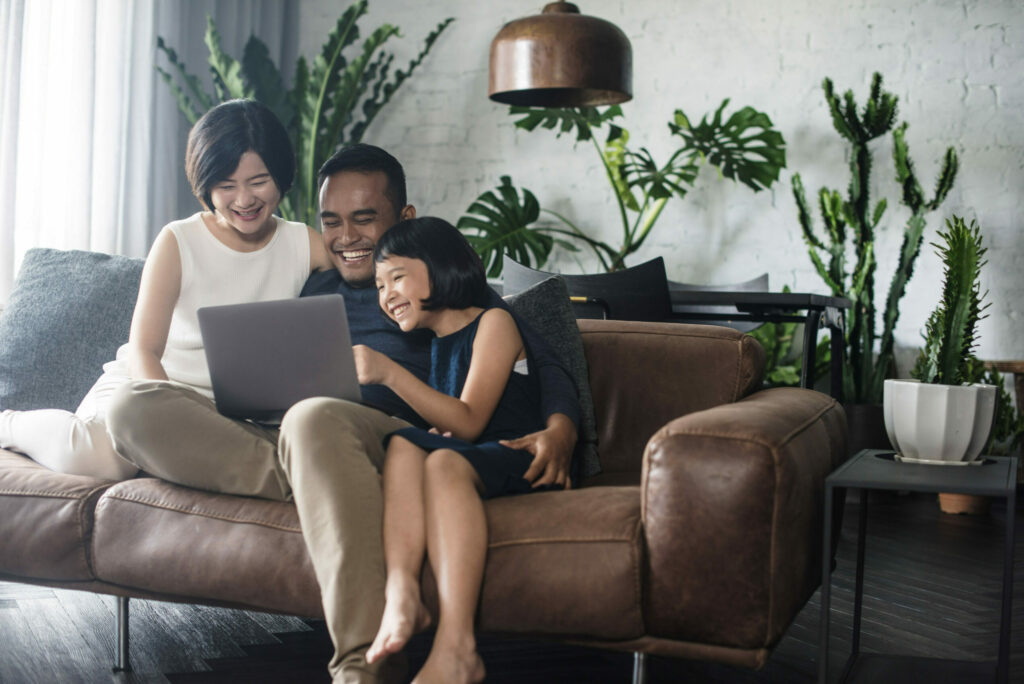 Couple heureux assis sur le sofa avec leur jeune fille, les yeux rivés sur un écran d’ordinateur portable dans une chambre avec plusieurs plantes