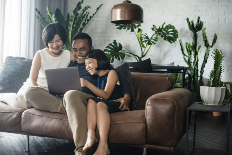Couple heureux assis sur le sofa avec leur jeune fille, les yeux rivés sur un écran d’ordinateur portable dans une chambre avec plusieurs plantes