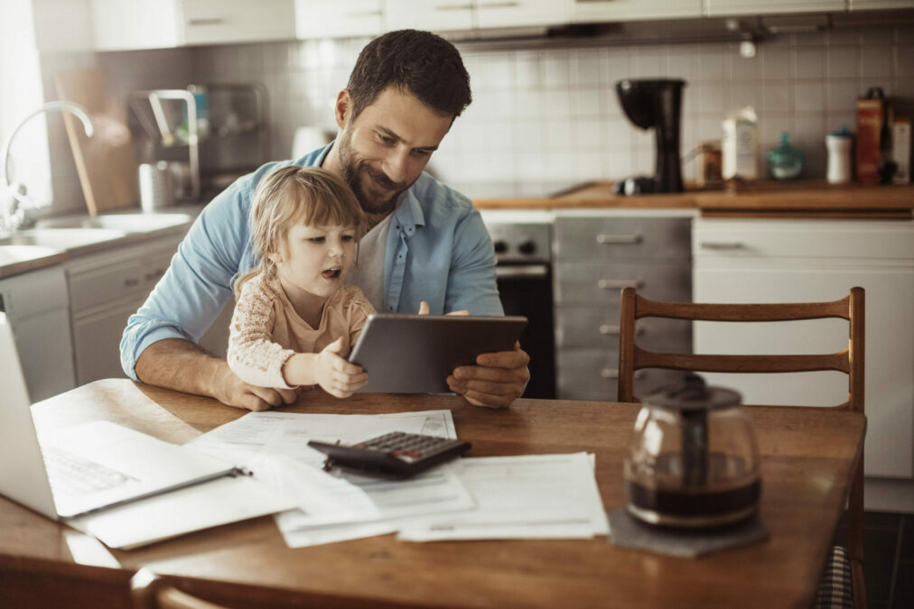Jeune fille assise sur les genoux de son père dans la cuisine pendant qu'il travaille à distance
