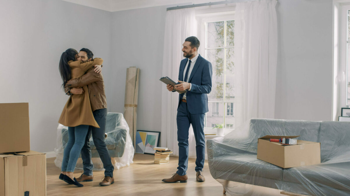 Un jeune couple s’embrasse dans leur nouvelle maison tandis que le courtier immobilier sourit