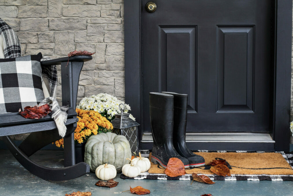 Gros plan de la porte d'entrée extérieure, noire, rehaussée d'une chaise à bascule, d'un coussin et d'une couverture à carreaux noirs et blancs, de citrouilles et de courges, de fleurs oranges et de bottes de pluie noires.