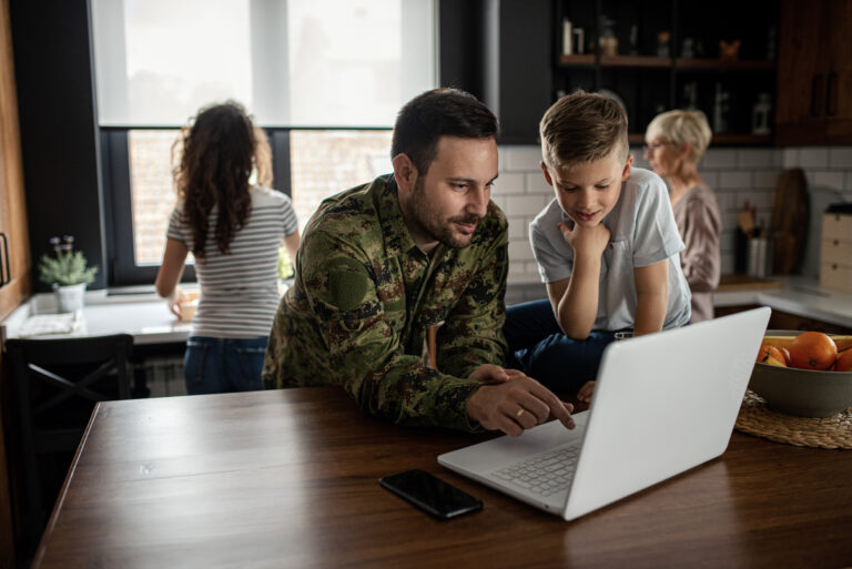 Un homme en uniforme militaire assis au comptoir de la cuisine avec un ordinateur portable et son jeune fils