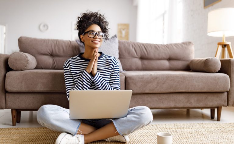 Une jeune femme avec une chemise rayée et des lunettes est assise devant le canapé avec un ordinateur portable