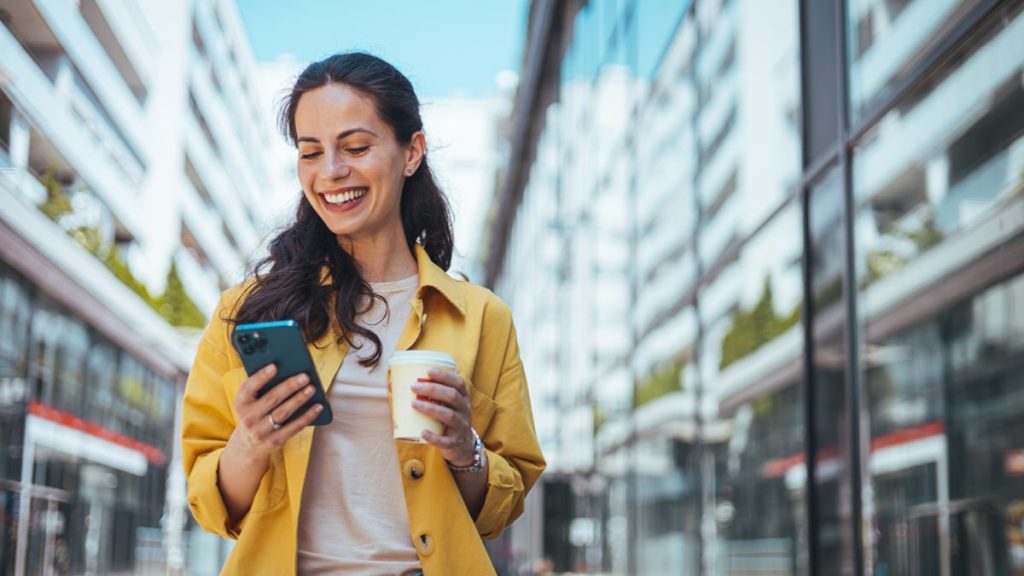 Femme en manteau jaune surfant sur Internet à l'aide d'un smartphone tout en buvant un café en plein air