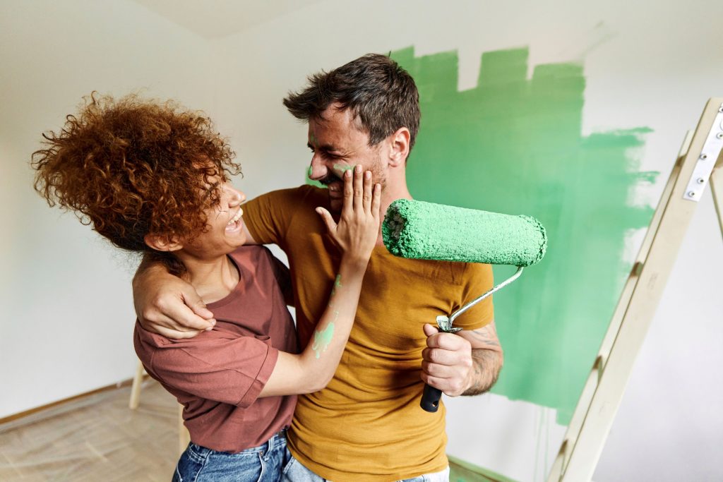 Un couple joyeux s'amuse pendant la rénovation d'une maison, tandis que la femme met de la peinture verte sur le visage de l'homme.