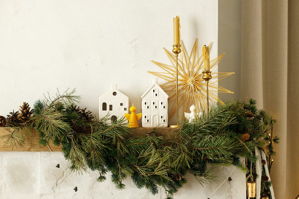Élégant manteau de cheminée décoré pour Noël avec branche de sapin, bougies, maisons en céramique, clochettes et ruban.