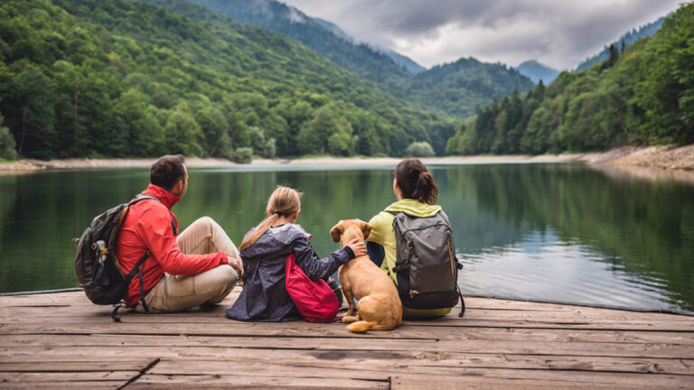 Une famille avec un petit chien jaune se repose sur un quai en admirant un lac et des montagnes brumeuses.