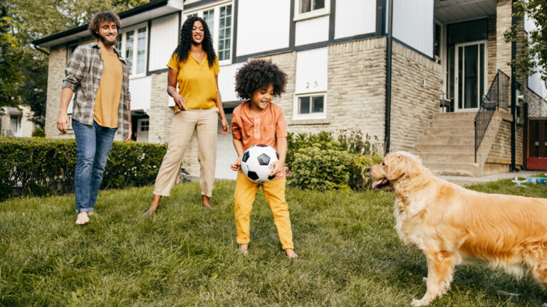 Une jeune famille jouant au ballon de foot sur la pelouse devant chez elle, en compagnie de son chien