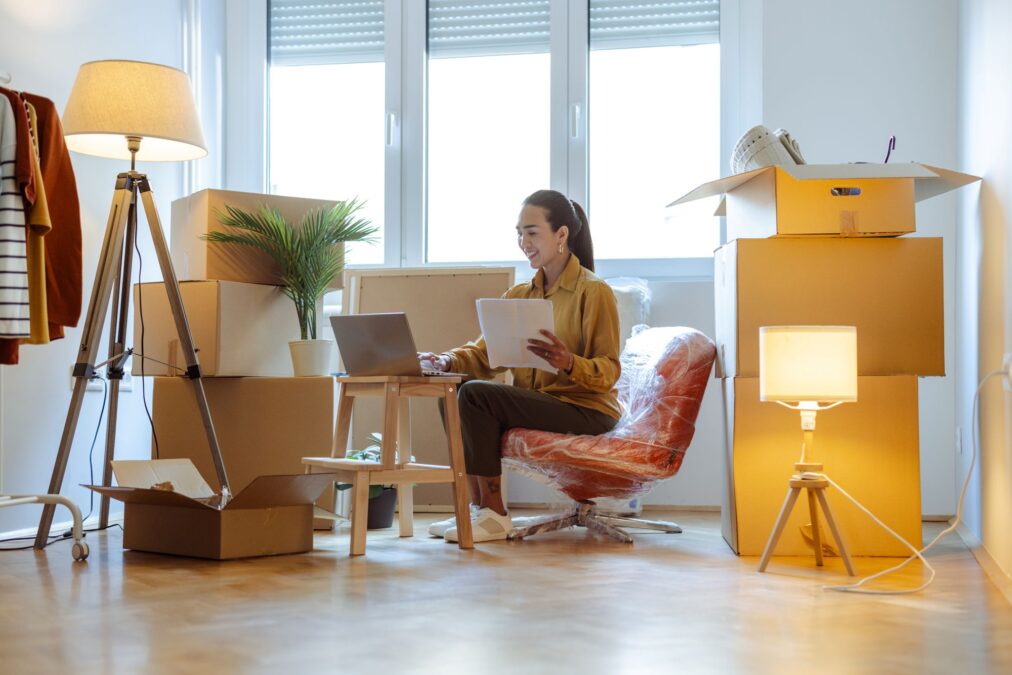 Une femme d'affaires effectuant plusieurs tâches à la fois sur son ordinateur portable, entourée de cartons dans sa nouvelle maison.