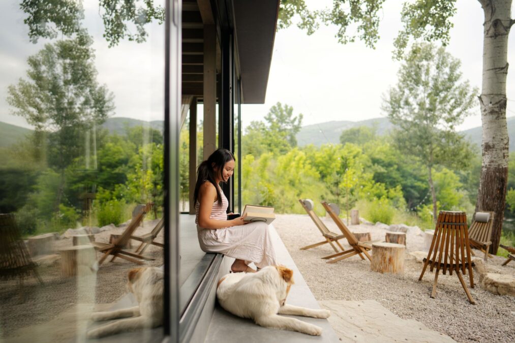 Photo d’une jeune femme en train de lire un livre sous la galerie de sa maison de montagne, en compagnie de son chien